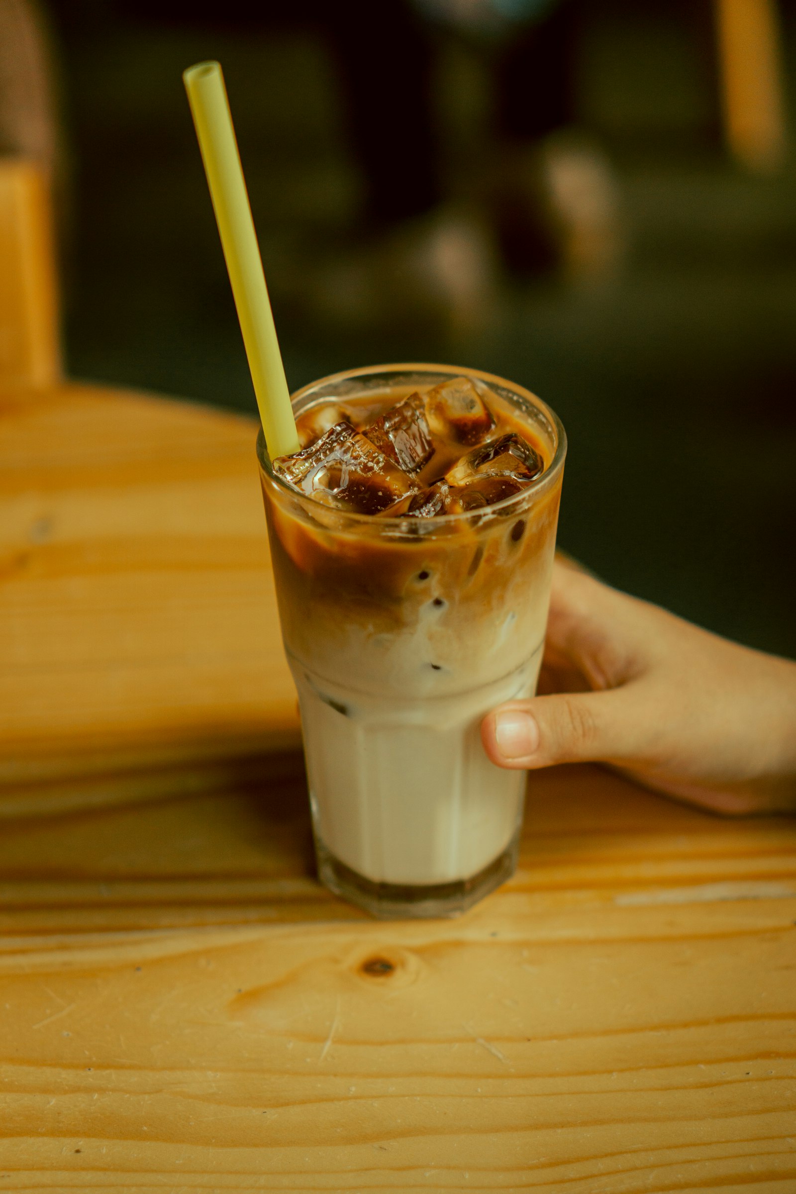 A hand resting beside a glass of iced milk tea with a straw on a wooden table